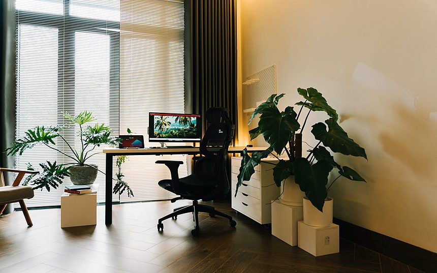 A home office corner with a desk, a large window and several indoor plants